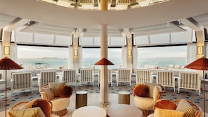 Un vaste espace intérieur aménagé avec de nombreux coins salon, de grandes fenêtres donnant sur l’océan et une colonne centrale.