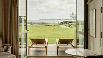 A room with a balcony featuring two wooden chairs and a small table, overlooking a grassy area with a wooden fence and a body of water in the distance.