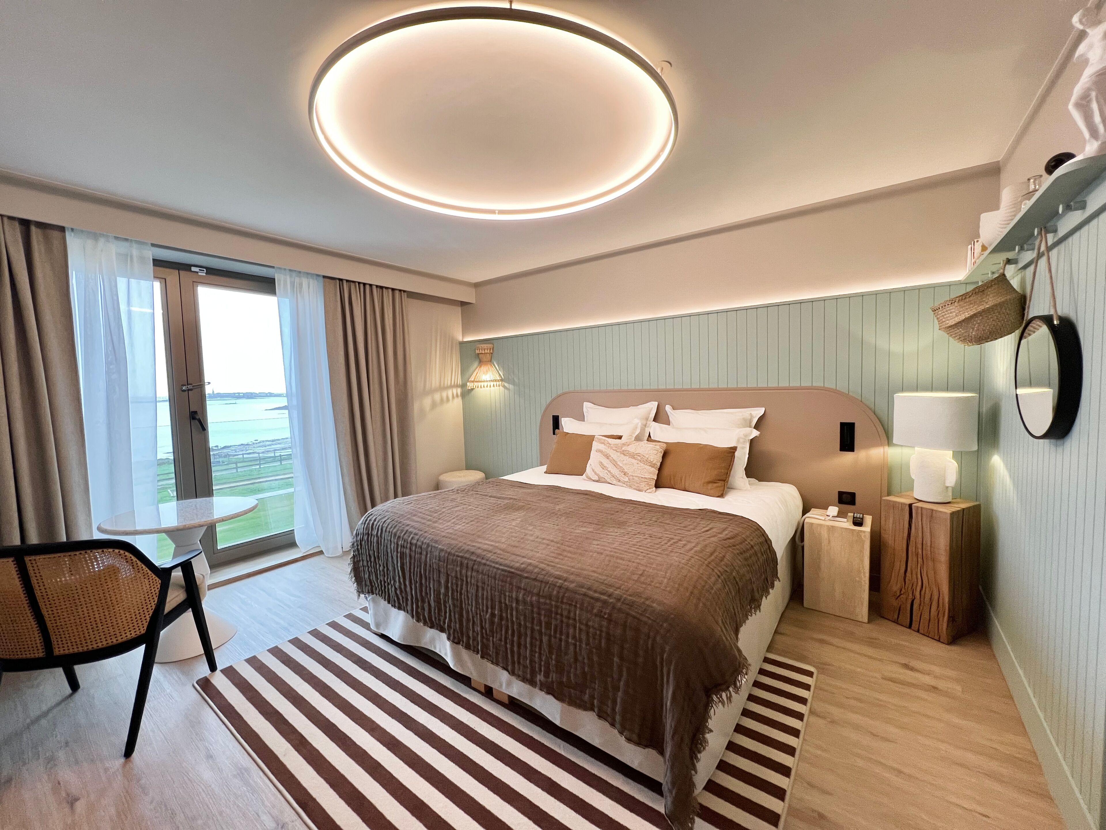 Habitación de hotel moderna con una cama grande, una lámpara de techo redonda, vistas al exterior, una mesita y una silla.