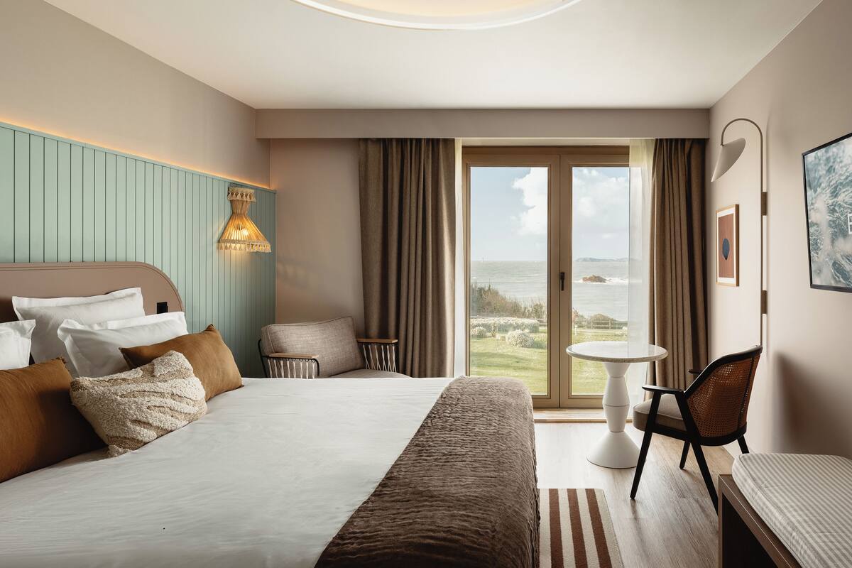 Une chambre d’hôtel avec un grand lit, une chaise, une petite table et une vue sur l’océan par une porte coulissante.
