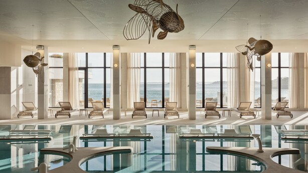 Une grande piscine intérieure dotée de plusieurs couloirs de nage, de chaises longues et de grandes fenêtres offrant une vue sur l’océan.