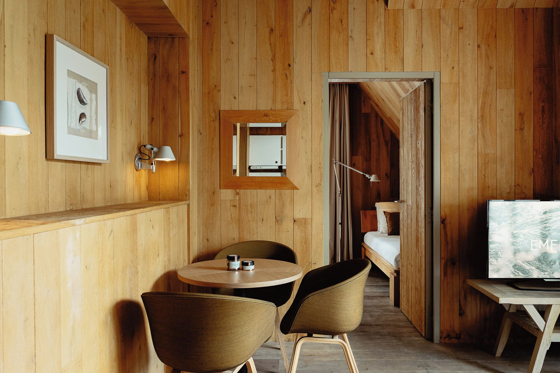 Een houten interieur met een ronde tafel, twee stoelen, een televisie en een ingelijst schilderij aan de muur.