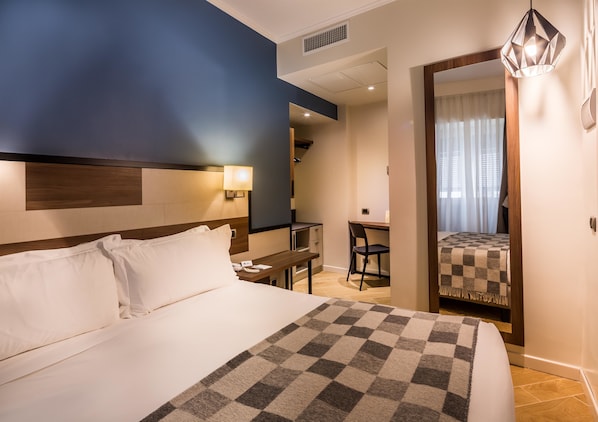 Une chambre d’hôtel avec un grand lit, un bureau et une chaise. Il y a un miroir au mur et une fenêtre avec des rideaux.
