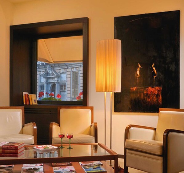 Une chambre d’hôtel avec deux fauteuils en cuir, une table basse en verre sur laquelle sont posés des magazines, une grande fenêtre donnant sur un immeuble, un grand lampadaire et un tableau accroché au mur.