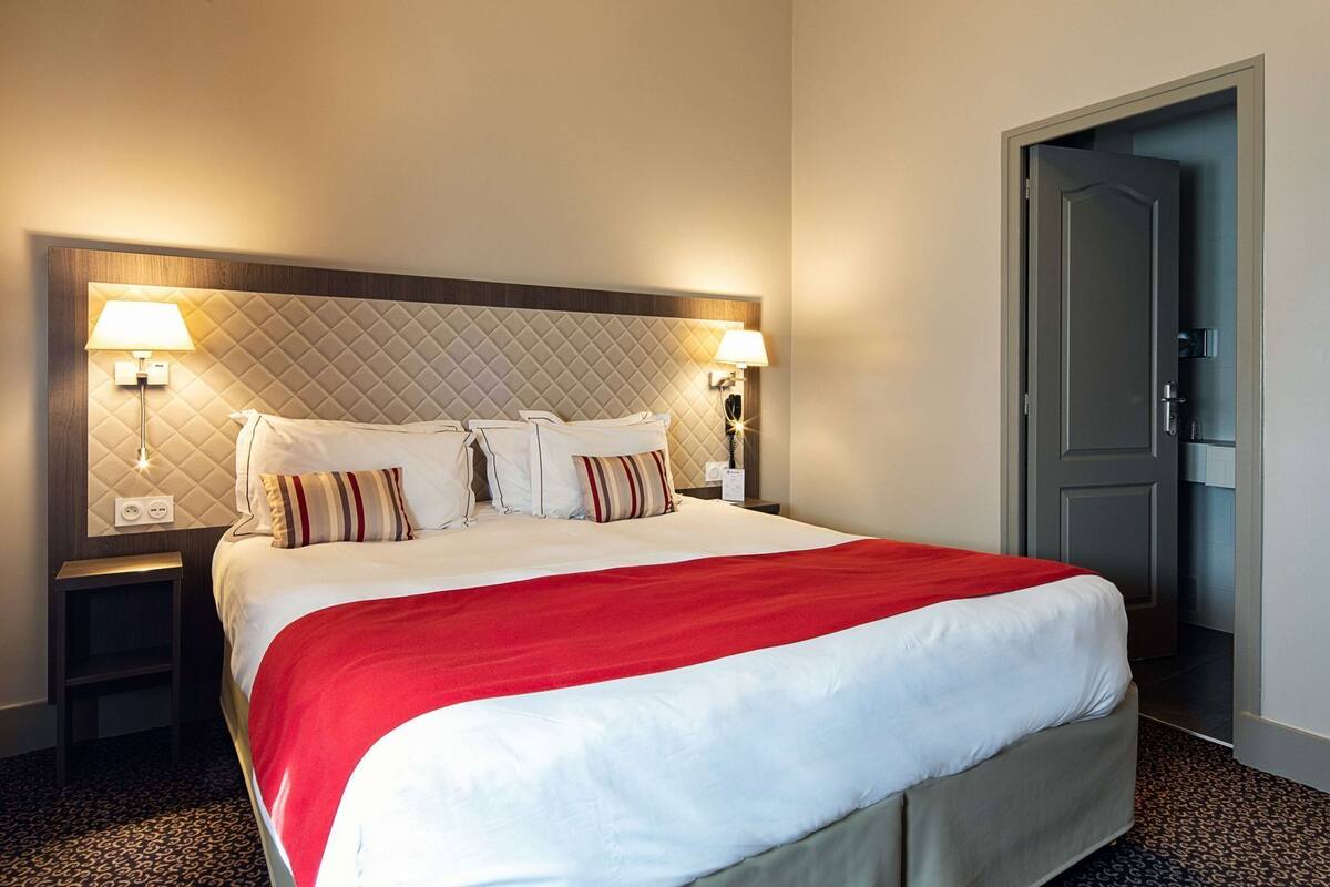 A hotel room with a large bed, two bedside tables with lamps, a door, and a patterned carpet.