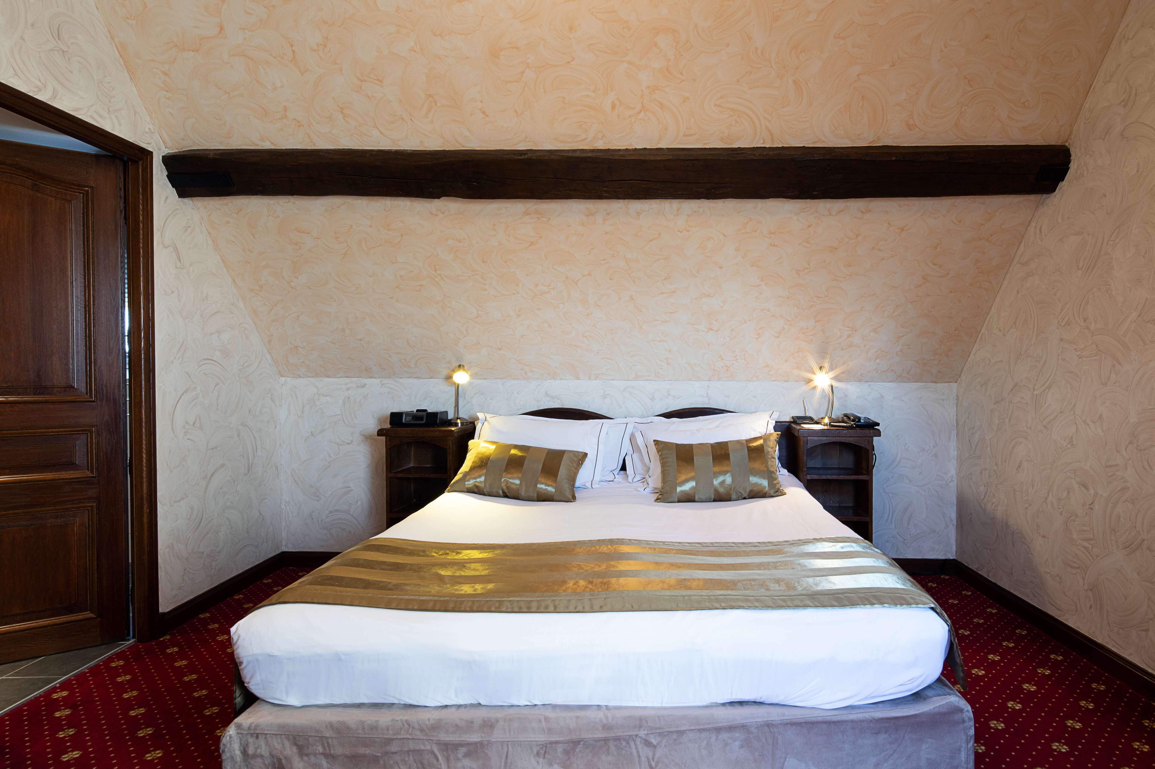 A neatly made bed with a striped blanket, two bedside lamps, and a wooden headboard in a room with patterned wallpaper and a red carpet.