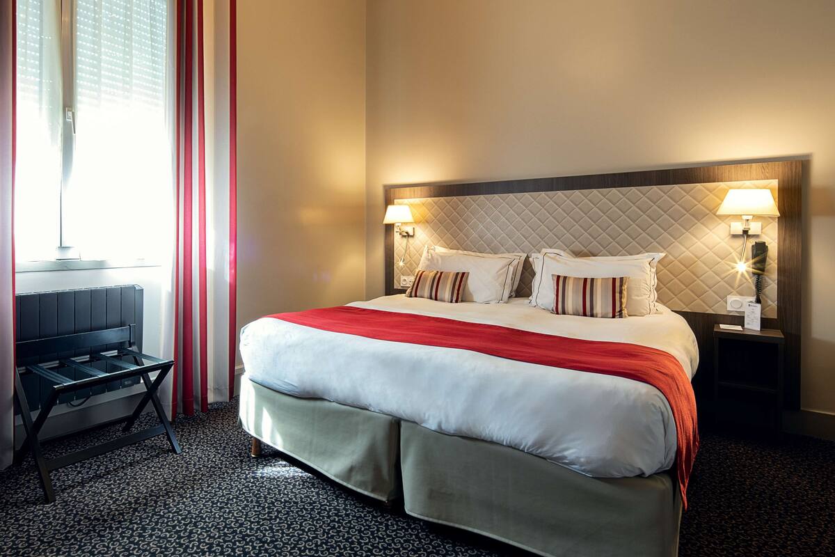 A hotel room with a bed, bedside tables, a desk, and a window with curtains.