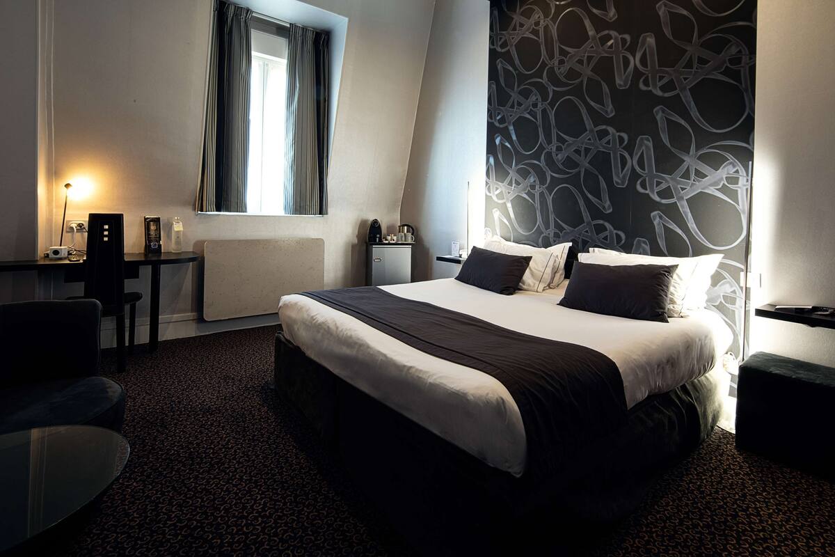 A modern hotel room with a large bed, a desk with a lamp, a small table, and a chair.