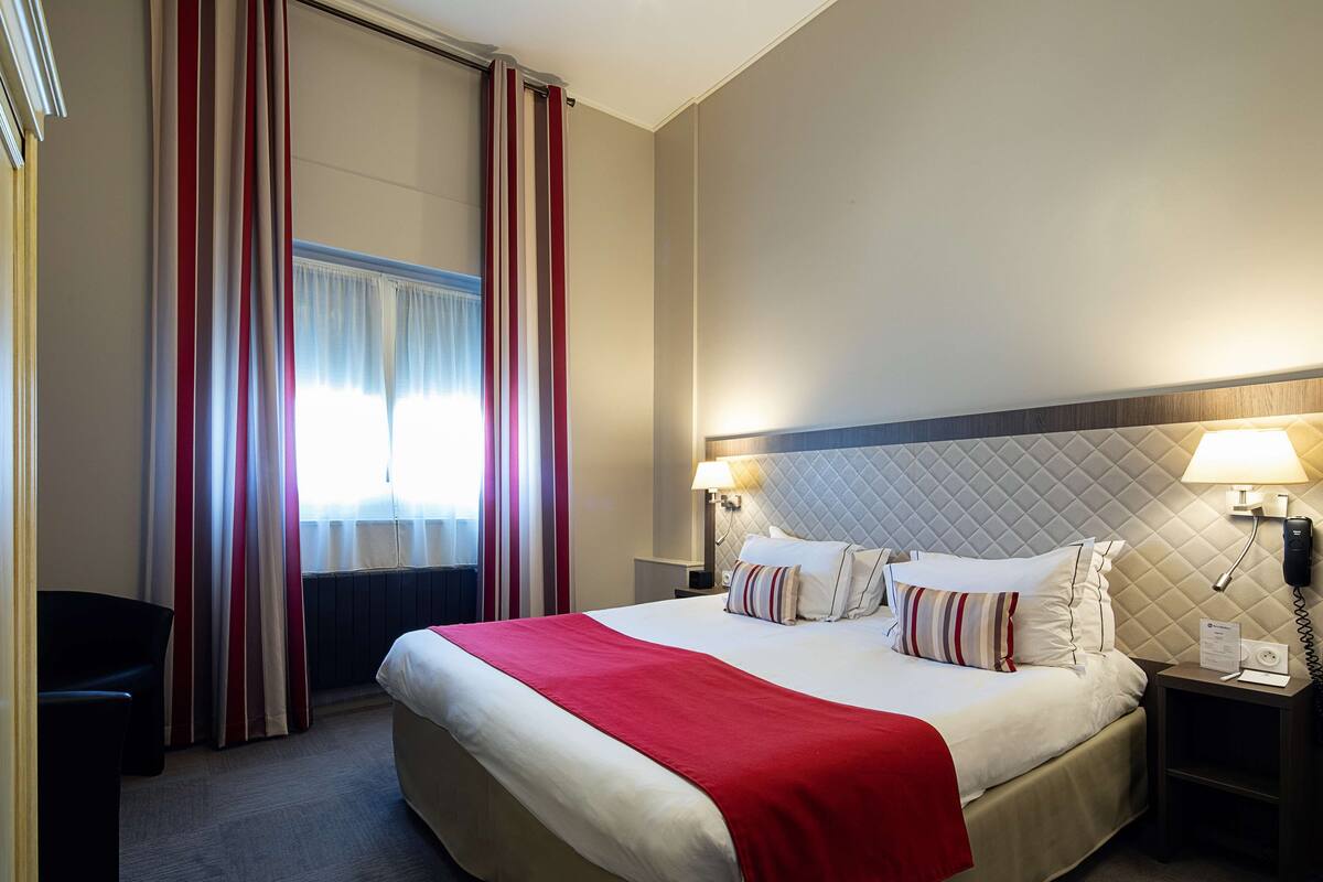 A hotel room with a large bed, two bedside lamps, a red throw blanket, and a view of the outdoors through curtains.