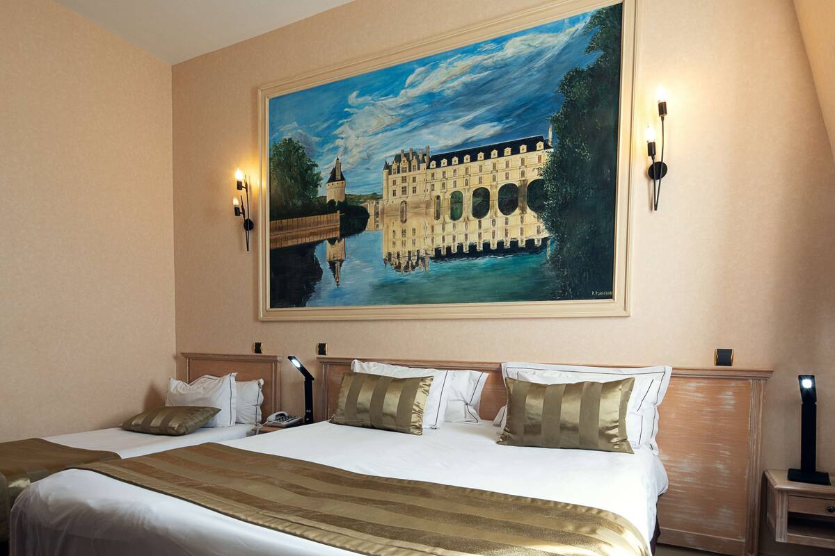 A hotel room with a large bed, two bedside tables, a painting of a castle on the wall, and two wall-mounted lamps.