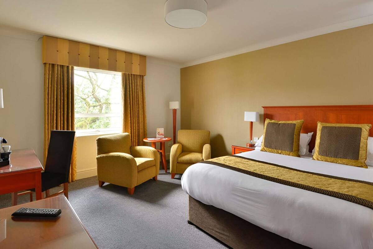 Habitación de hotel con cama, dos sillones, un escritorio con un control remoto, una lámpara y una ventana con cortinas.