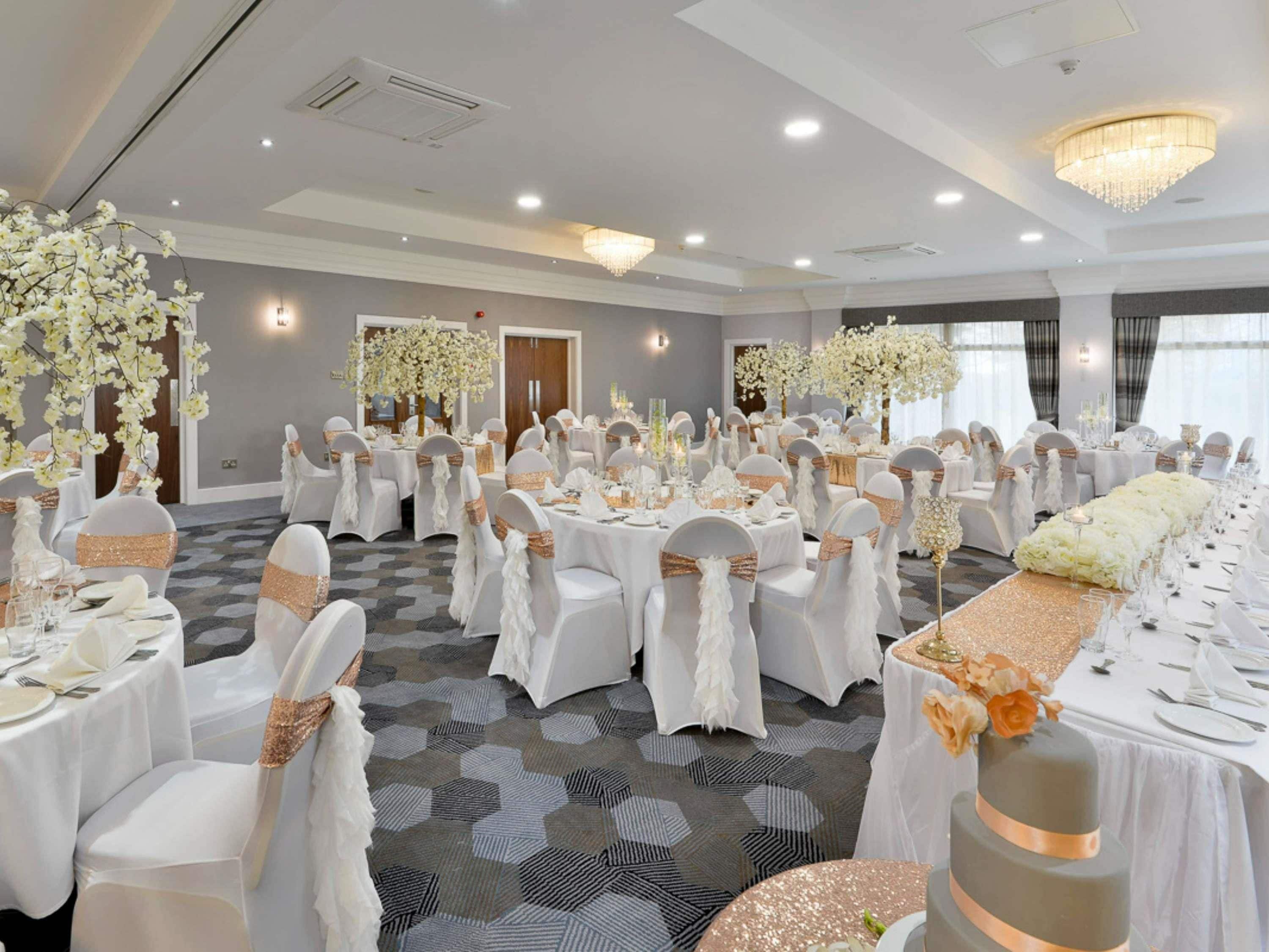 Een feestzaal met ronde tafels, witte tafellakens en stoelen met witte hoezen en gouden linten.