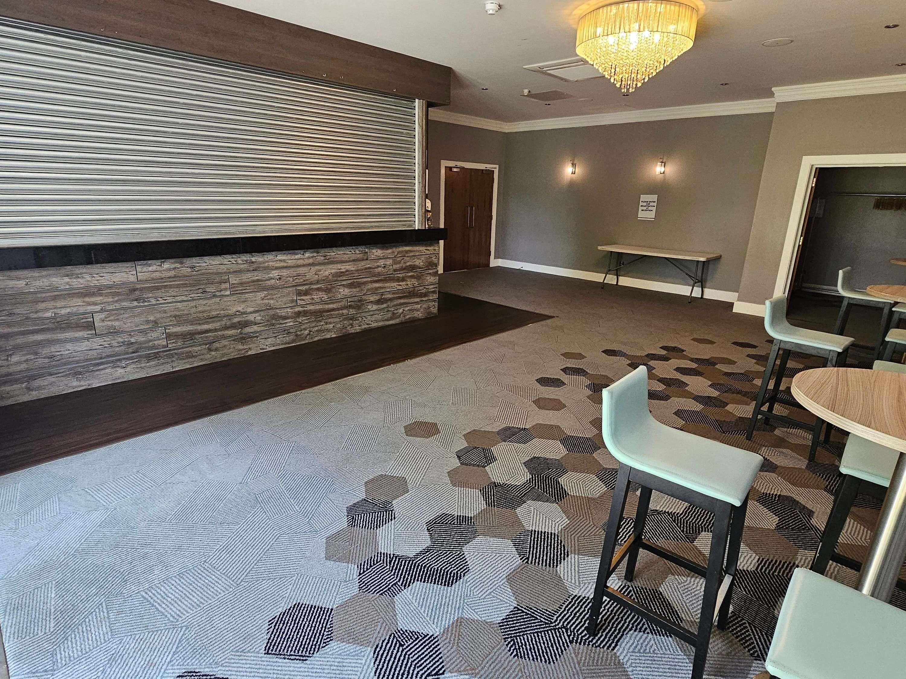 Un vestíbulo de hotel con alfombra estampada, un panel de madera decorativo, una araña y una zona de estar con mesas y sillas.