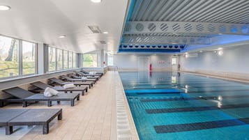 A swimming pool with sun loungers and a tiled floor, surrounded by windows.