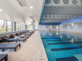 Una piscina con tumbonas y suelo de baldosas, rodeada de ventanas.