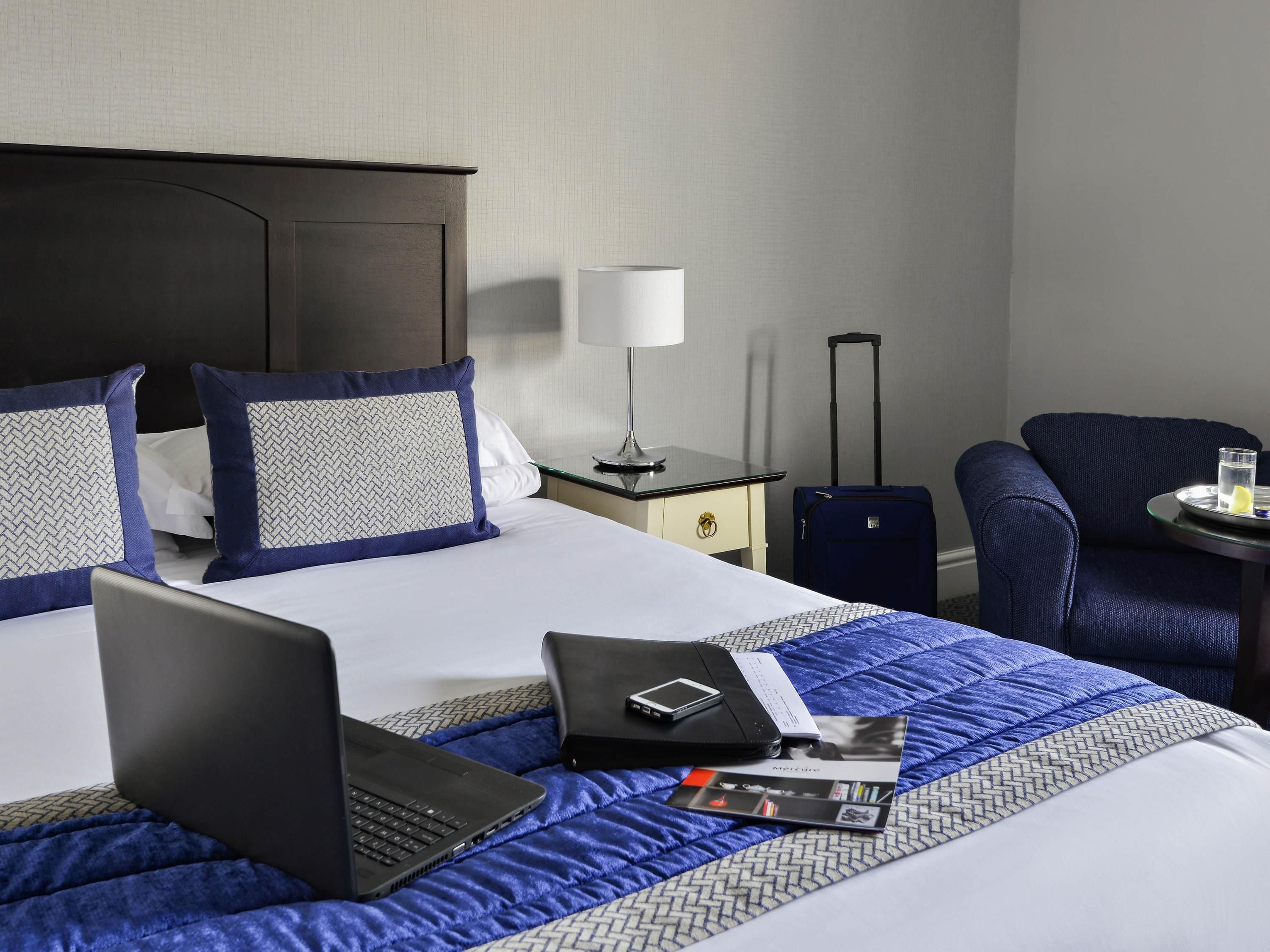 A hotel room with a bed, a desk, a chair, a laptop, a suitcase, and a lamp.