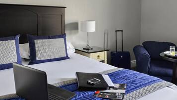 A hotel room with a bed, a desk, a chair, a laptop, a suitcase, and a lamp.