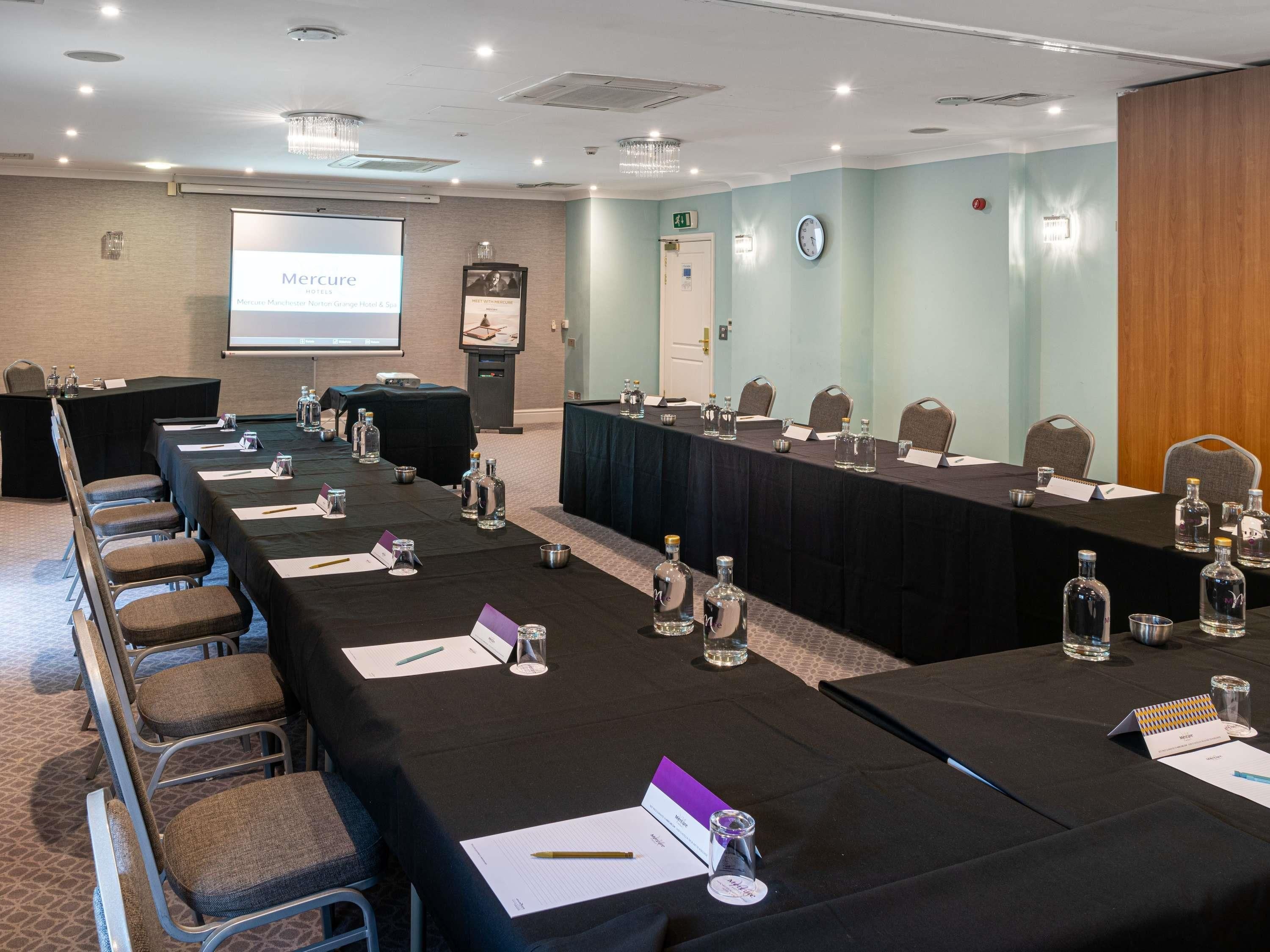 Una sala de conferències amb taules llargues, cadires, plaques de noms, ampolles d'aigua i una pantalla de projector que mostra una presentació.