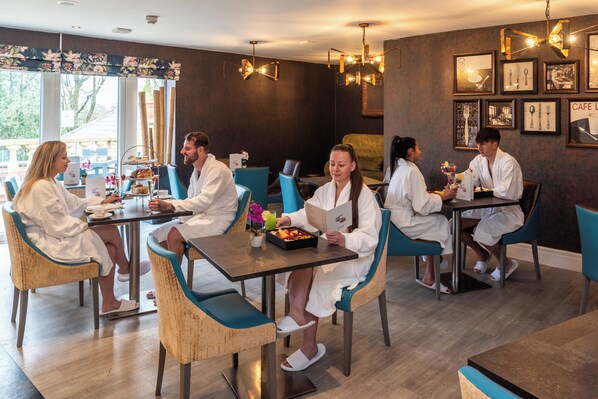 Un grupo de personas vestidas de blanco sentadas en una mesa con una bandeja de comida y una bebida, en una habitación con piso de madera y sillas azules.