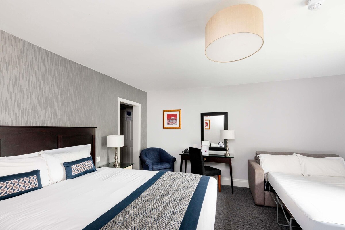 Une chambre d’hôtel avec un grand lit, un bureau avec une chaise, une lampe de bureau, une chaise et une table de chevet.