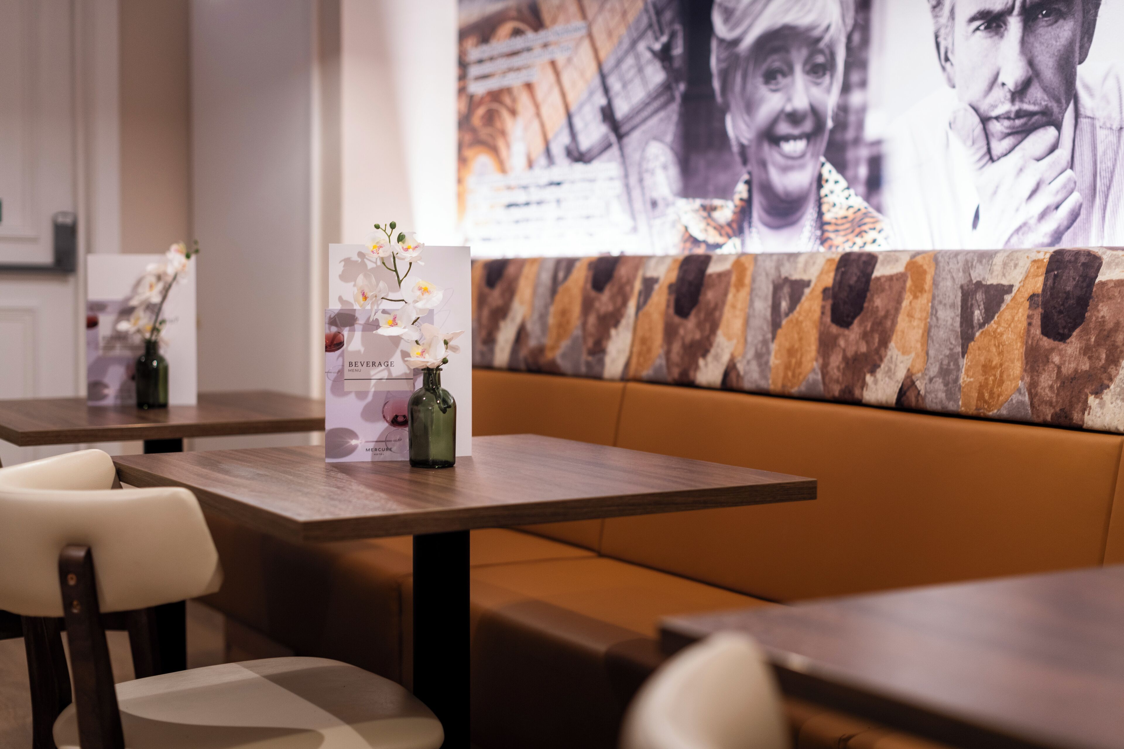 A restaurant interior with wooden tables and cushioned booths, a vase with flowers, and a wall with a large photograph.