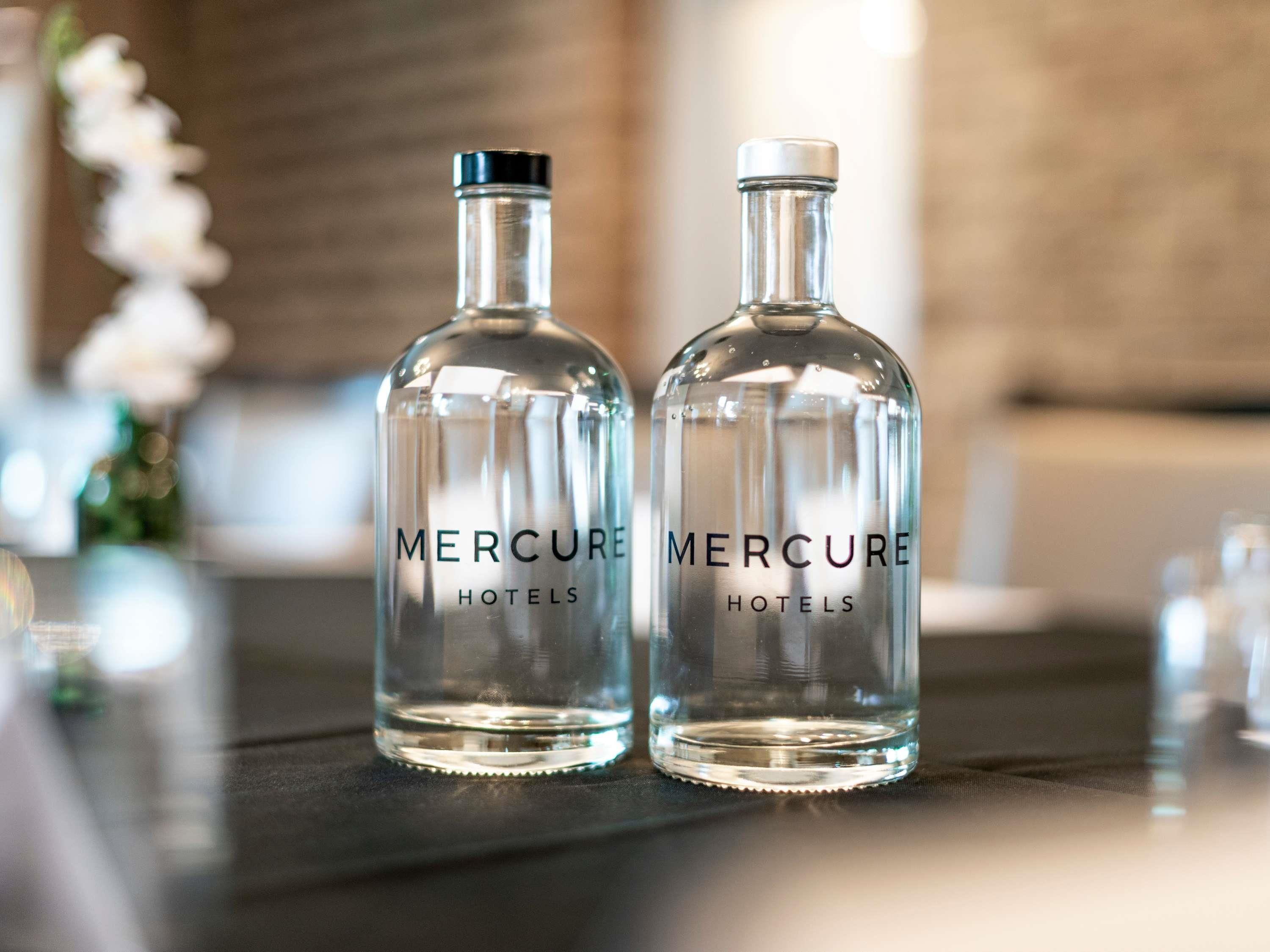 Deux bouteilles en verre transparent, portant l’étiquette « Mercure Hotels », sont posées sur une surface sombre.