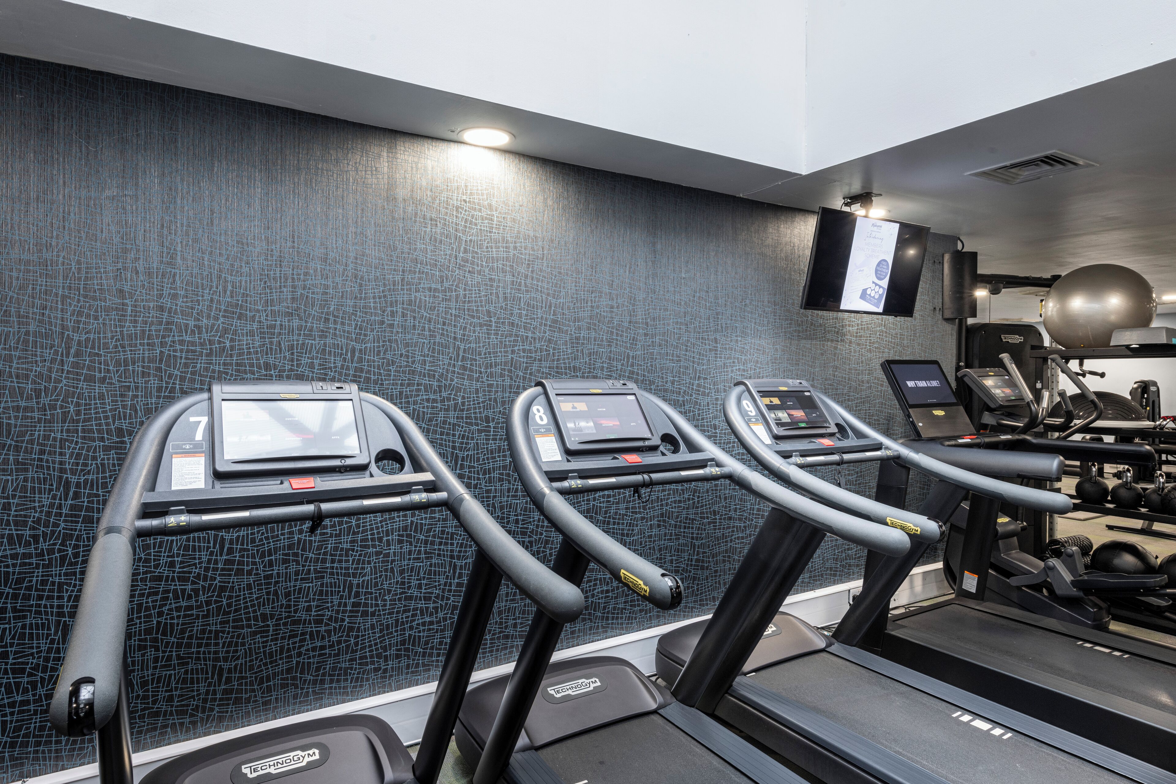 Tres cintas de correr en un gimnasio con una pared texturizada y un televisor montado.