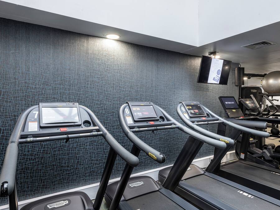 Tres cintas de correr en un gimnasio con una pared texturizada y un televisor montado.