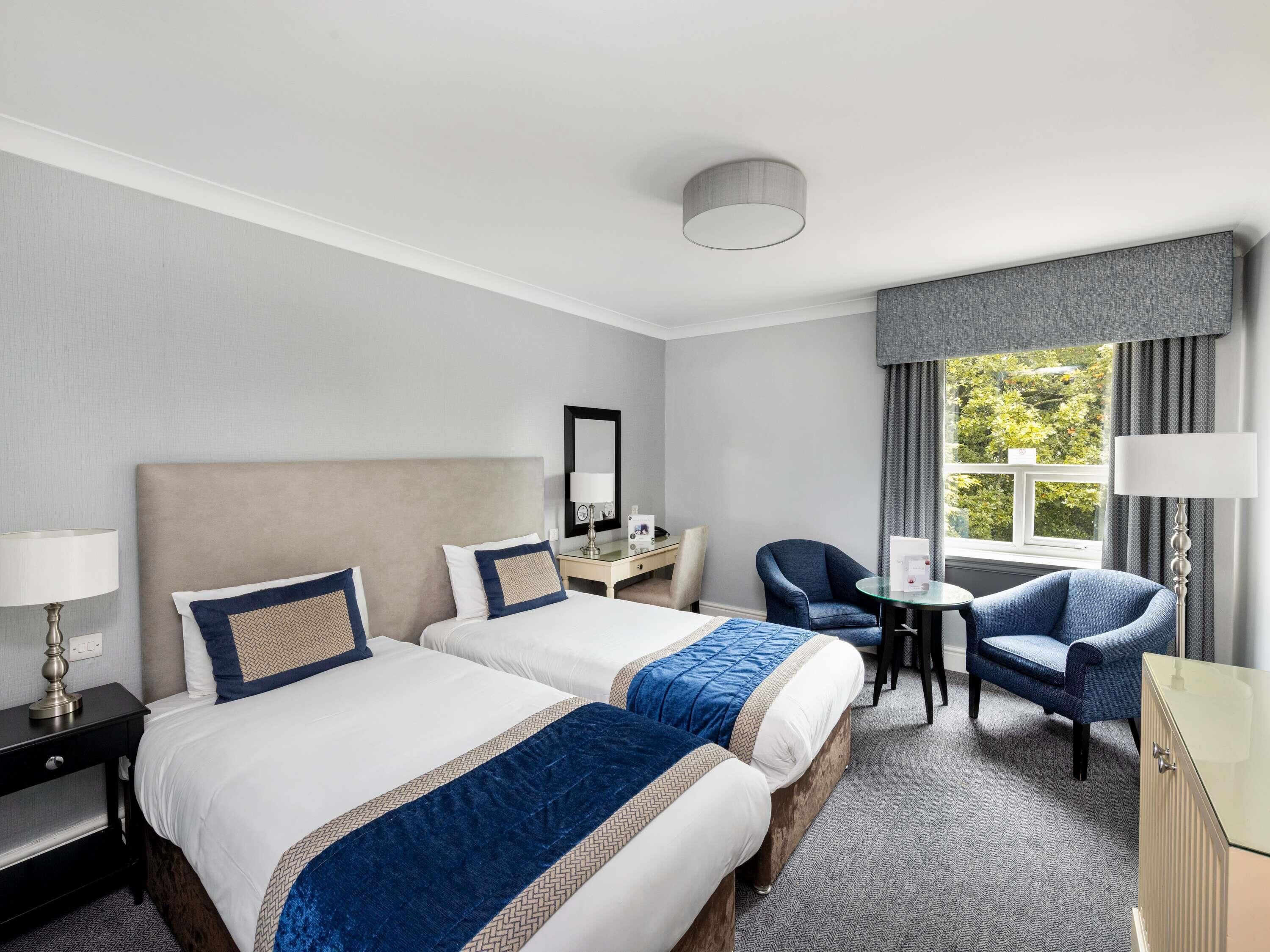 Hotellihuone, jossa on kaksi sänkyä, työpöytä, tuoli, lamppu ja näkymä vihreälle alueelle.