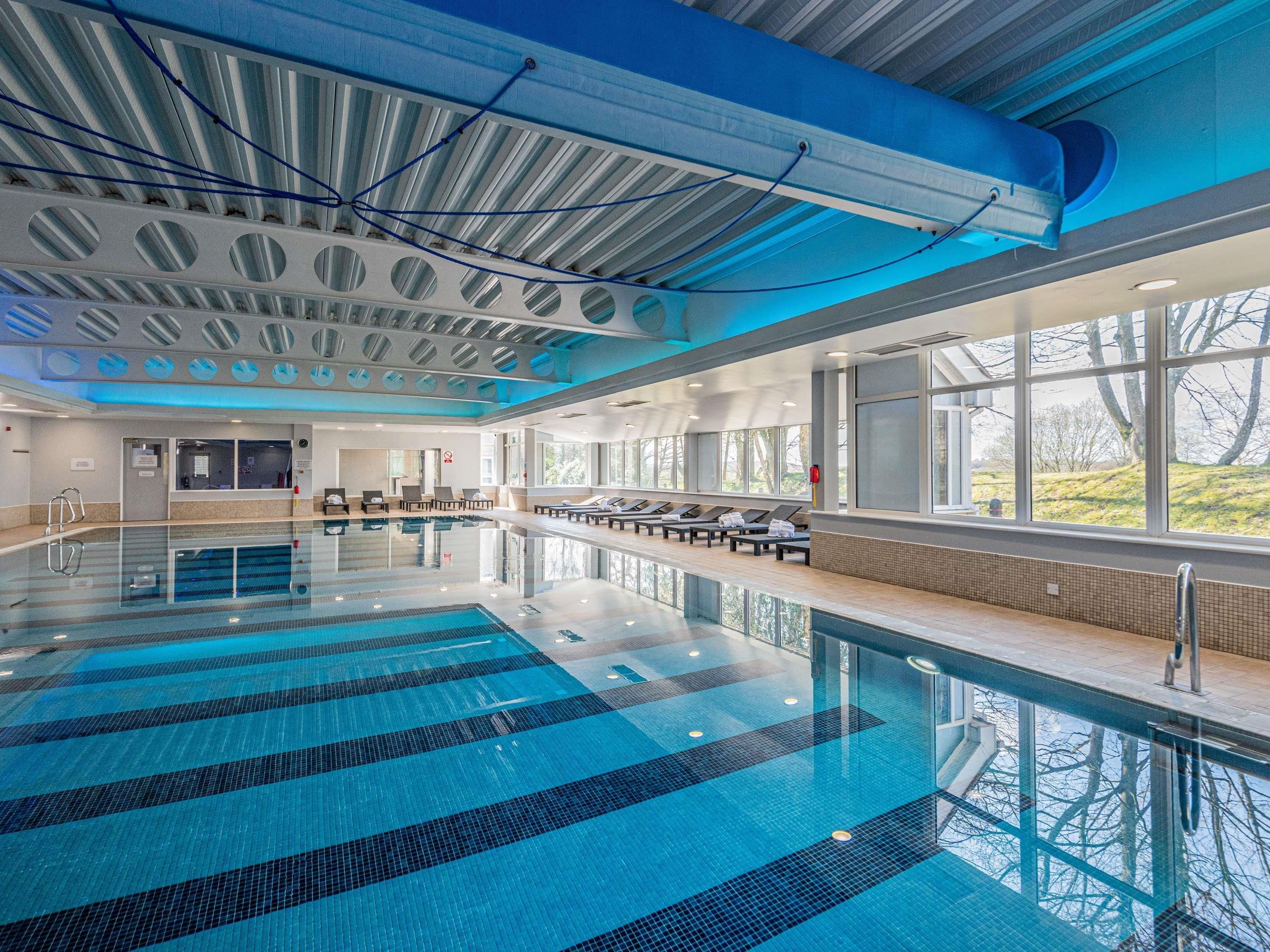 A large indoor swimming pool with clear blue water, sunlit windows, and lounge chairs along the side.