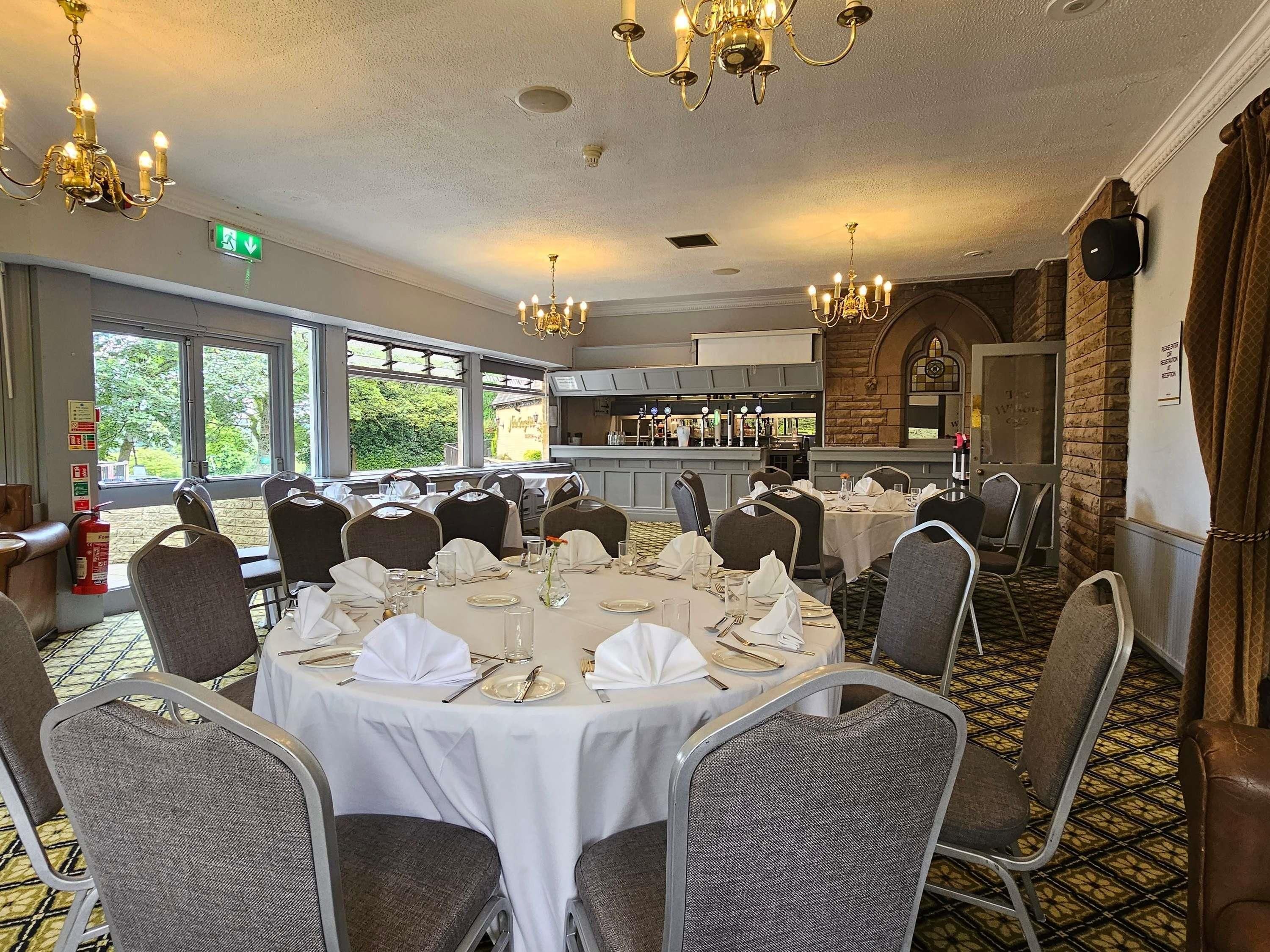 Un salón de banquetes con mesas redondas preparadas para comer, una zona de cocina y ventanas grandes que ofrecen vistas a zonas verdes.