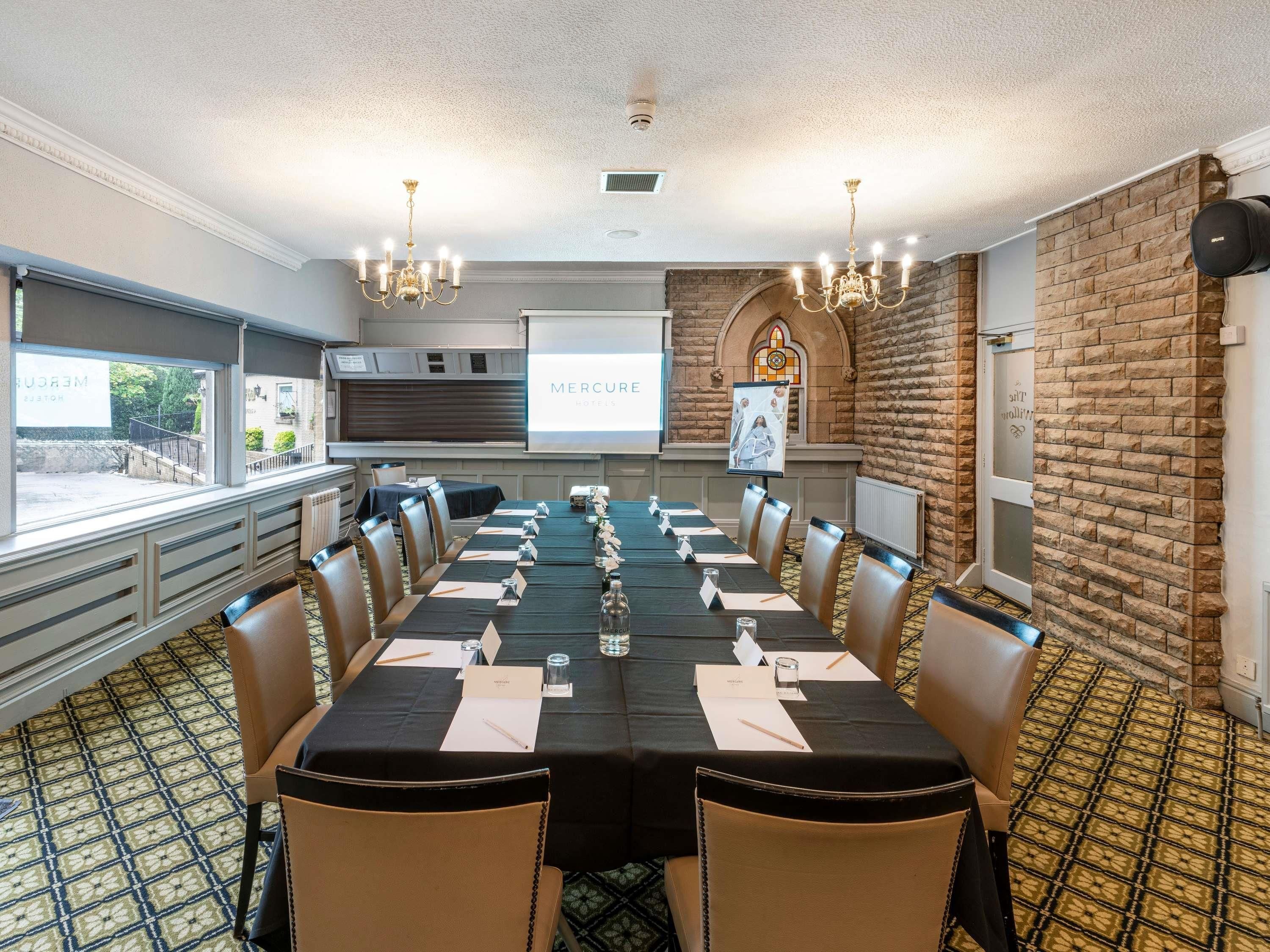 Un salón de conferencias con una larga mesa, sillas, una pantalla de proyector y una pared de ladrillo.