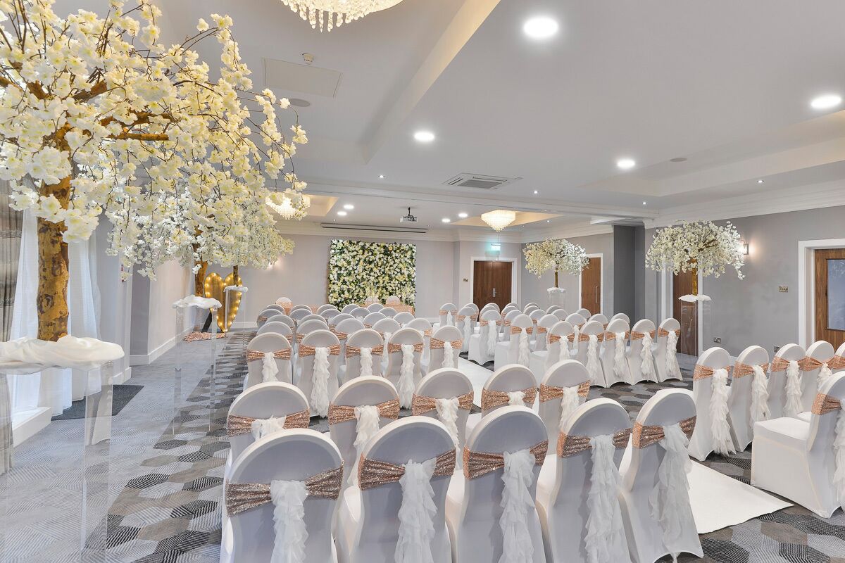 Un salón de banquetes con filas de sillas blancas, un arreglo floral grande y una araña.