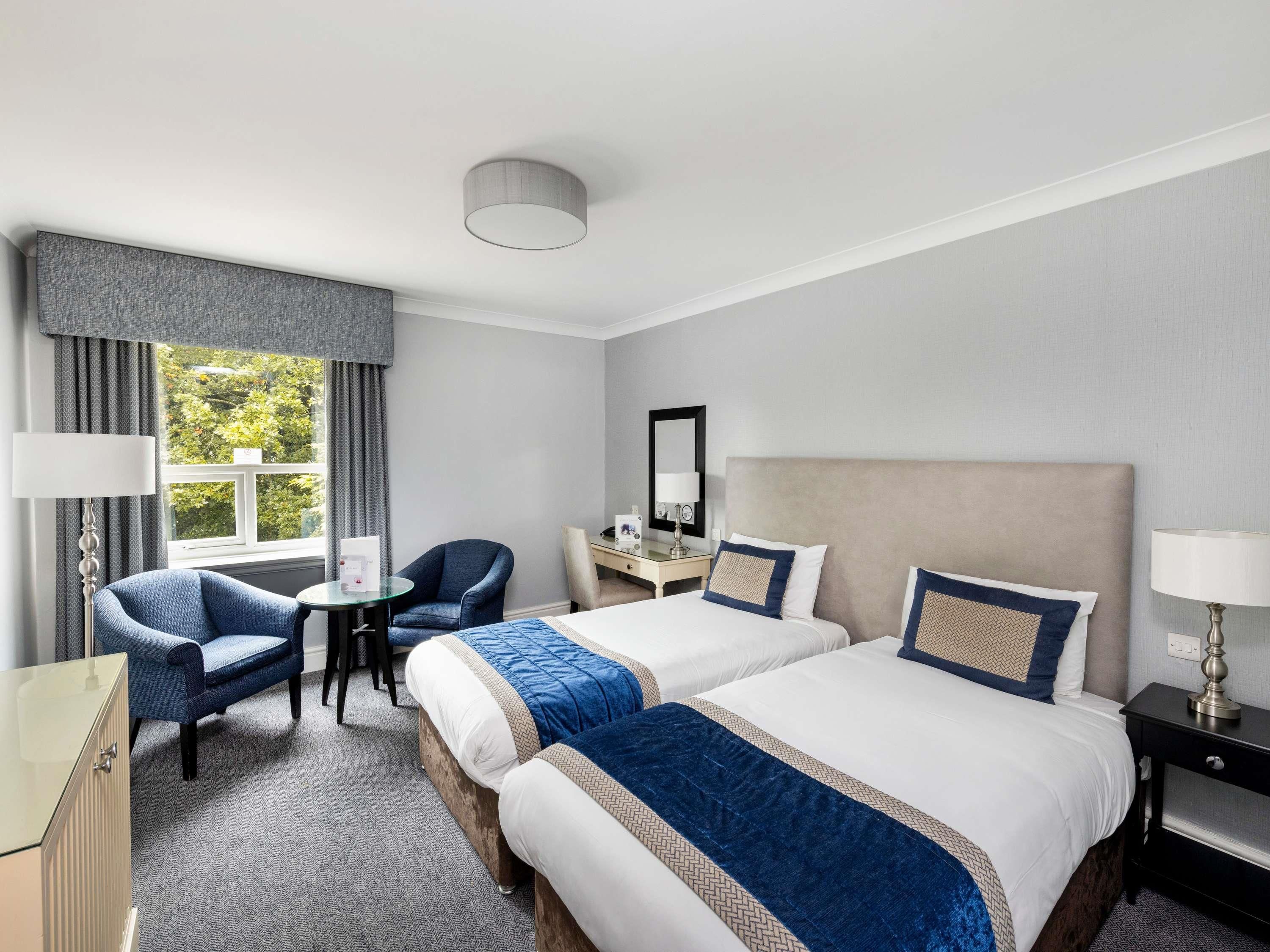 Habitación de hotel con dos camas, un escritorio, una silla, una lámpara y una ventana con cortinas.