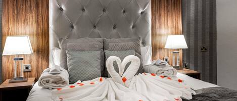 A hotel room with a tufted headboard, two bedside lamps, a bed with a white and gray duvet, and a red flower arrangement on the bed.