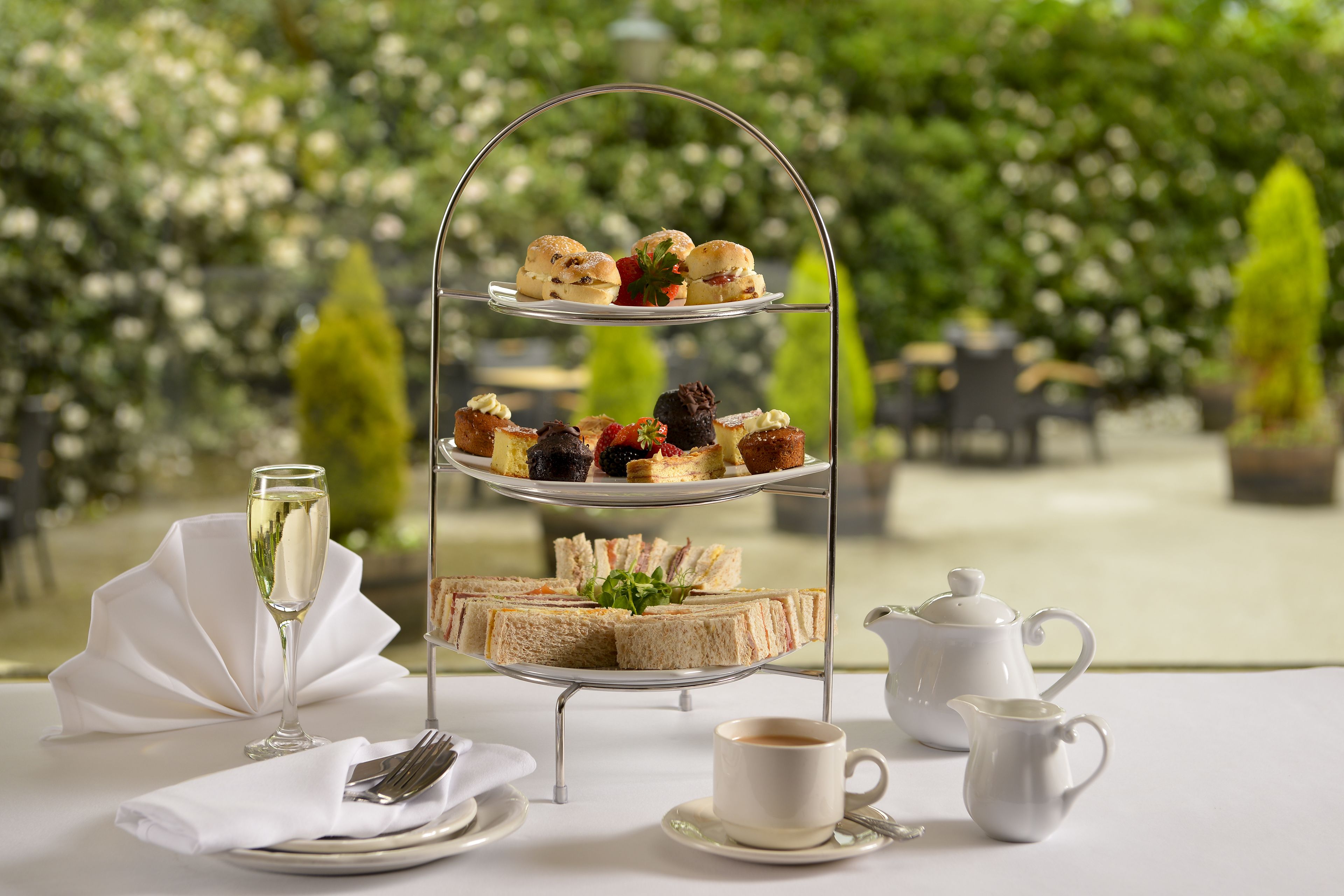 A three-tiered dessert stand with various pastries and sandwiches, a glass of champagne, a cup of coffee, and a teapot on a table with a white tablecloth.