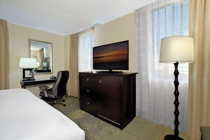 Quarto de hotel com cama, escrivaninha com luminária, televisão, cômoda e luminária.