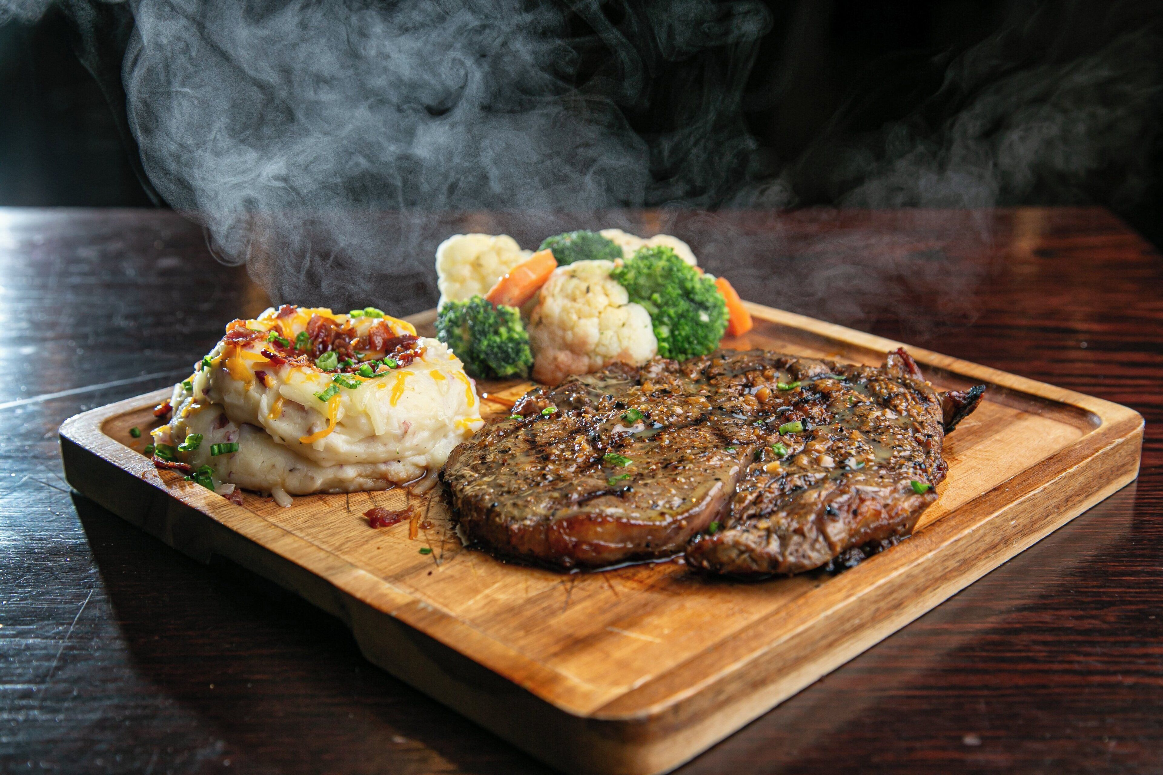 A steak with mashed potatoes and steamed vegetables on a wooden board.