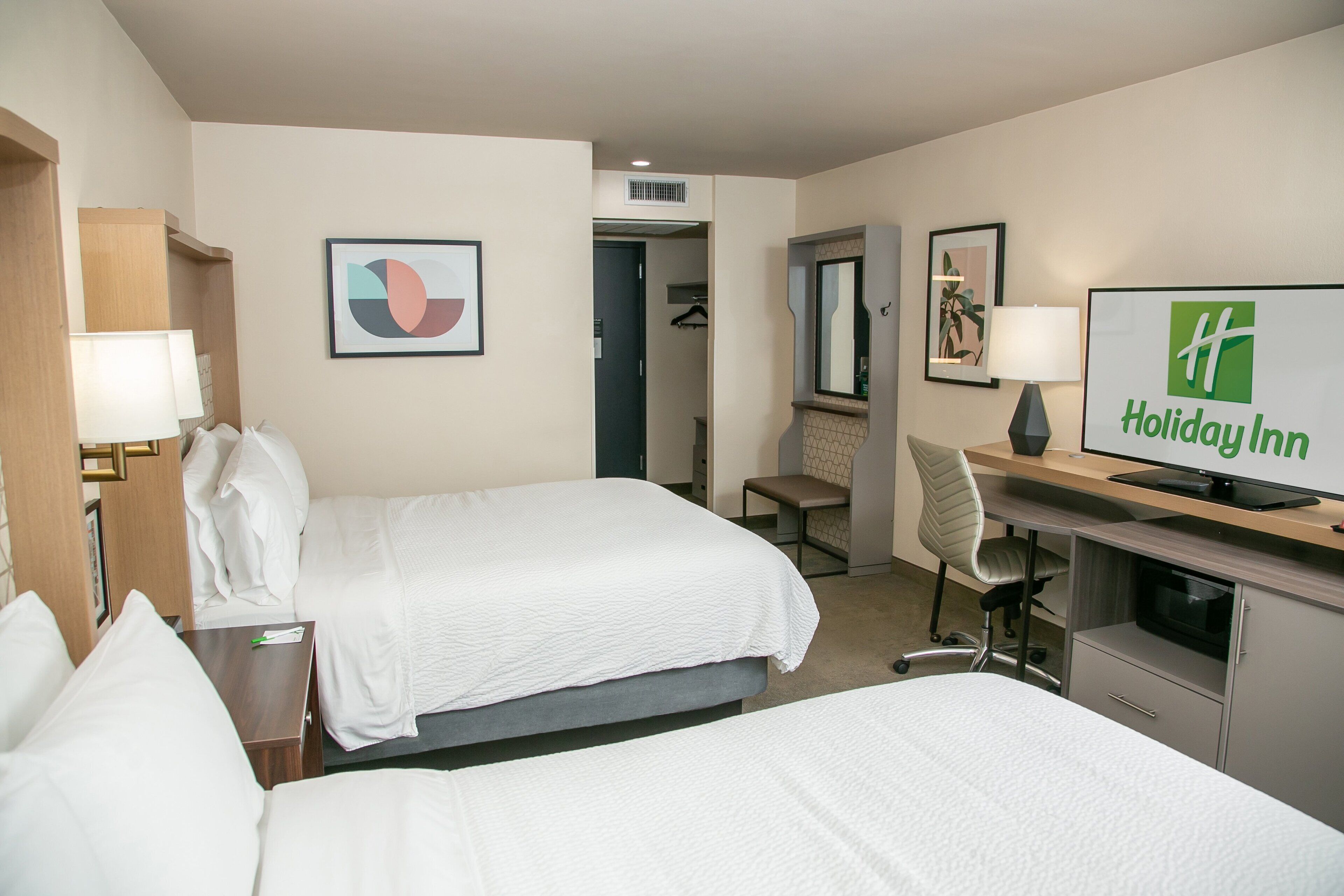 A hotel room with two beds, a desk with a computer, a chair, and a TV displaying the Holiday Inn logo.