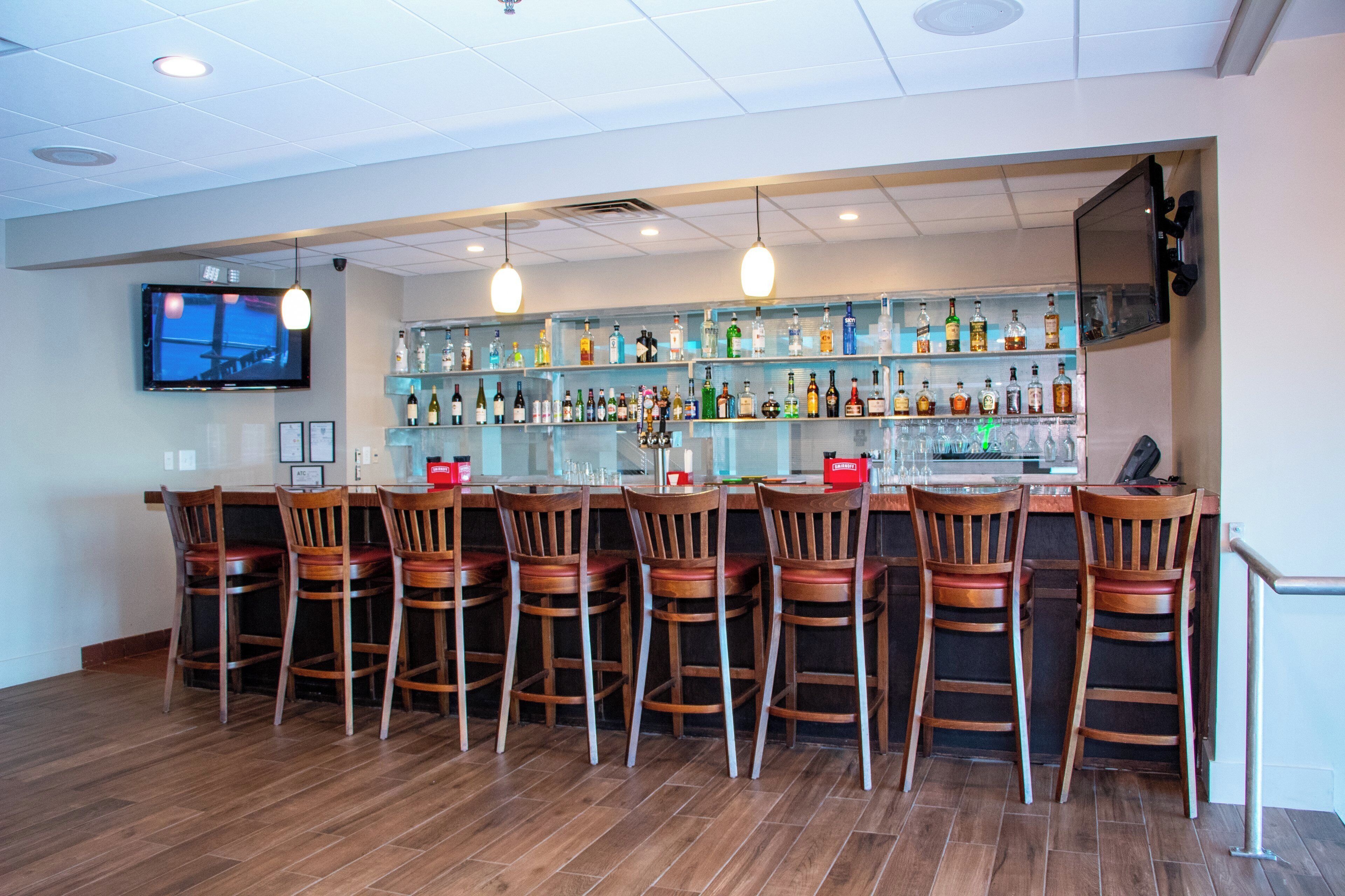 A bar area with multiple wooden bar stools, a variety of liquor bottles on shelves, and a flat-screen TV mounted on the wall.