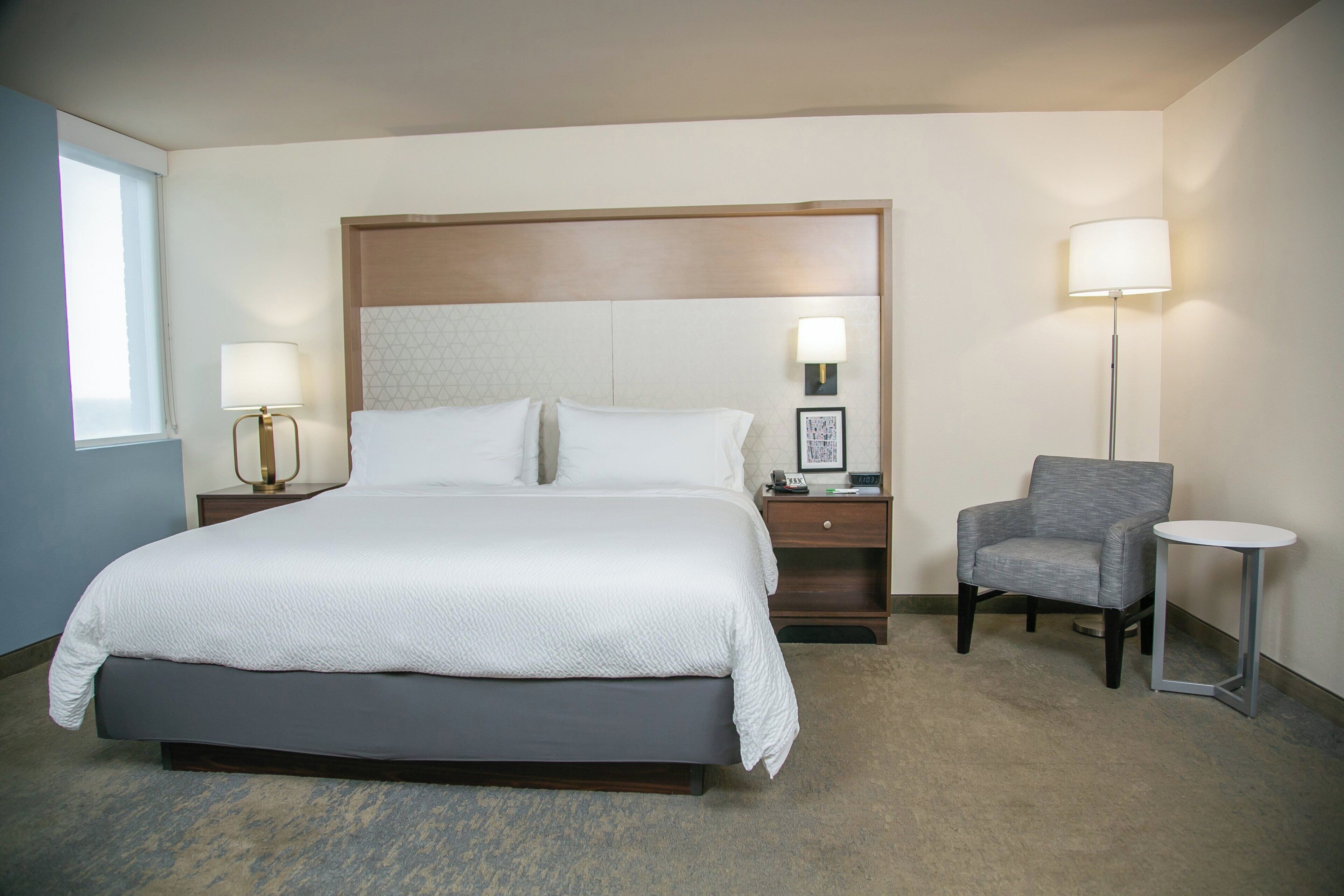 A hotel room with a large bed, two bedside tables with lamps, a gray armchair, and a small side table.