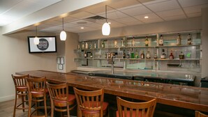 Zona de bar con una barra de madera larga, varios taburetes y estantes llenos de botellas.