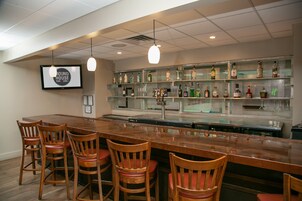 Zona de bar con una barra de madera larga, varios taburetes y estantes llenos de botellas.