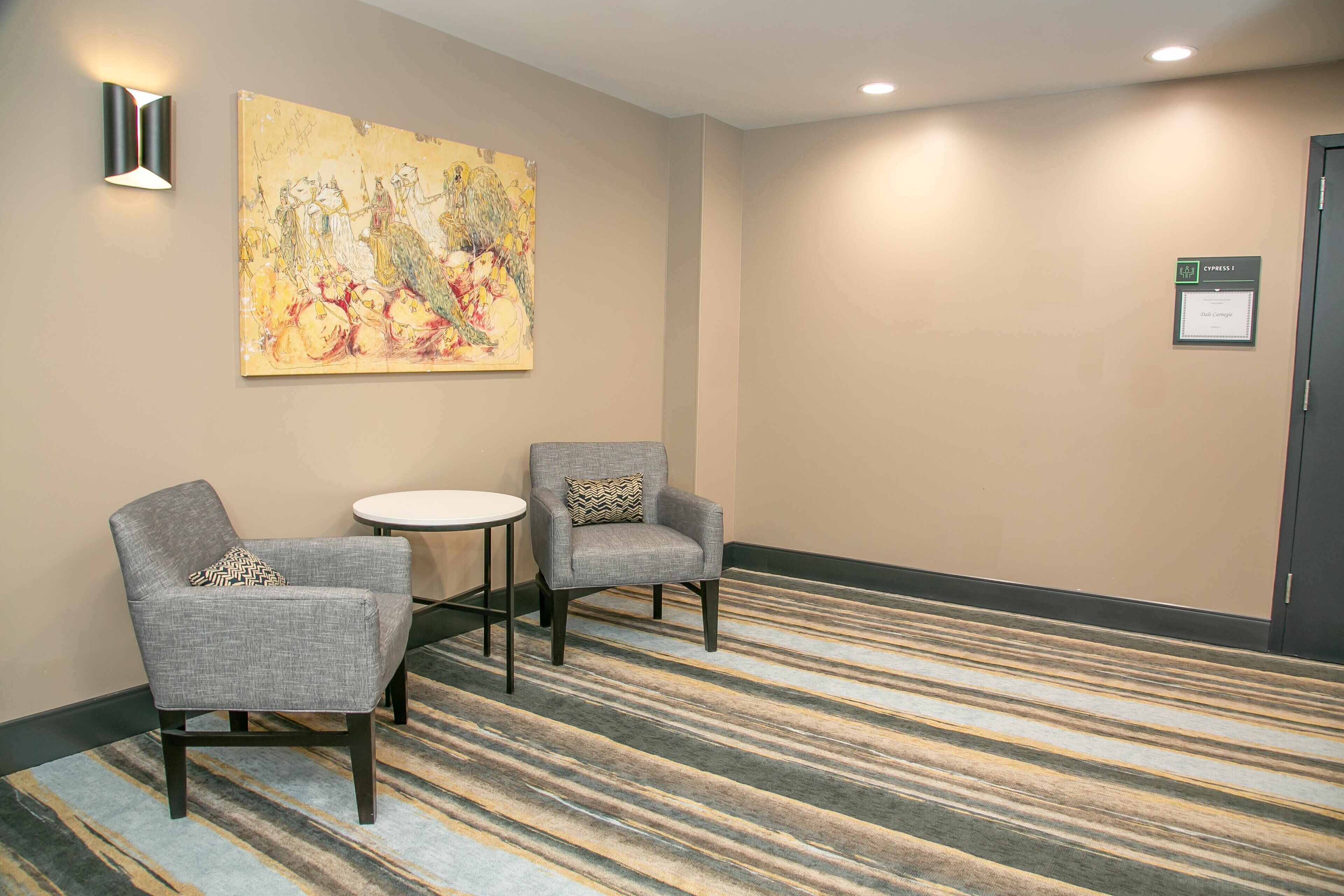 A hotel room with two grey armchairs, a small round table, a large abstract painting on the wall, and a striped carpet.