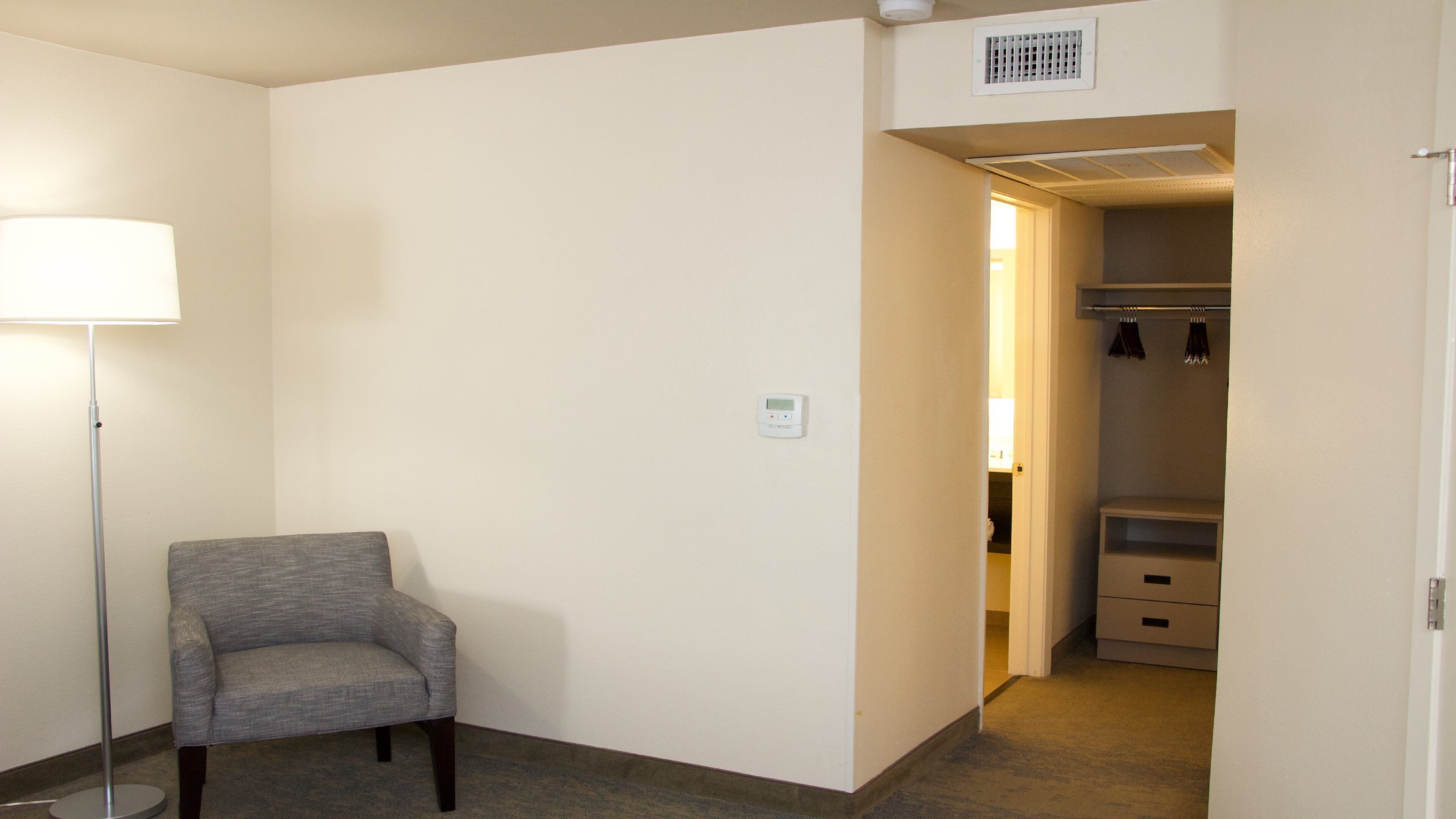 A hotel room with a grey armchair, a standing floor lamp, a closet with hanging clothes, and a visible thermostat on the wall.