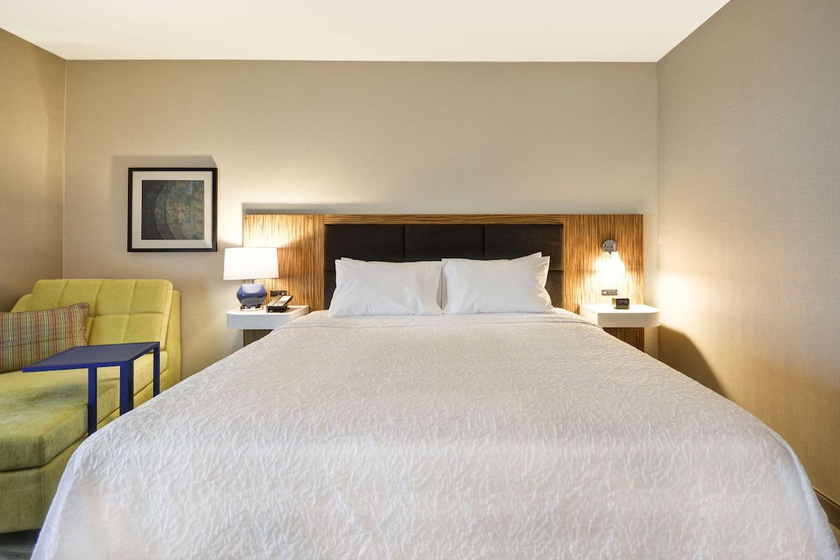 A hotel room with a large bed, a nightstand with a lamp, a small table, a green armchair, and a framed picture on the wall.