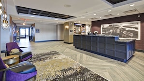 Een moderne hotellobby met een receptiebalie, een zithoek en een groot wandkunstwerk.