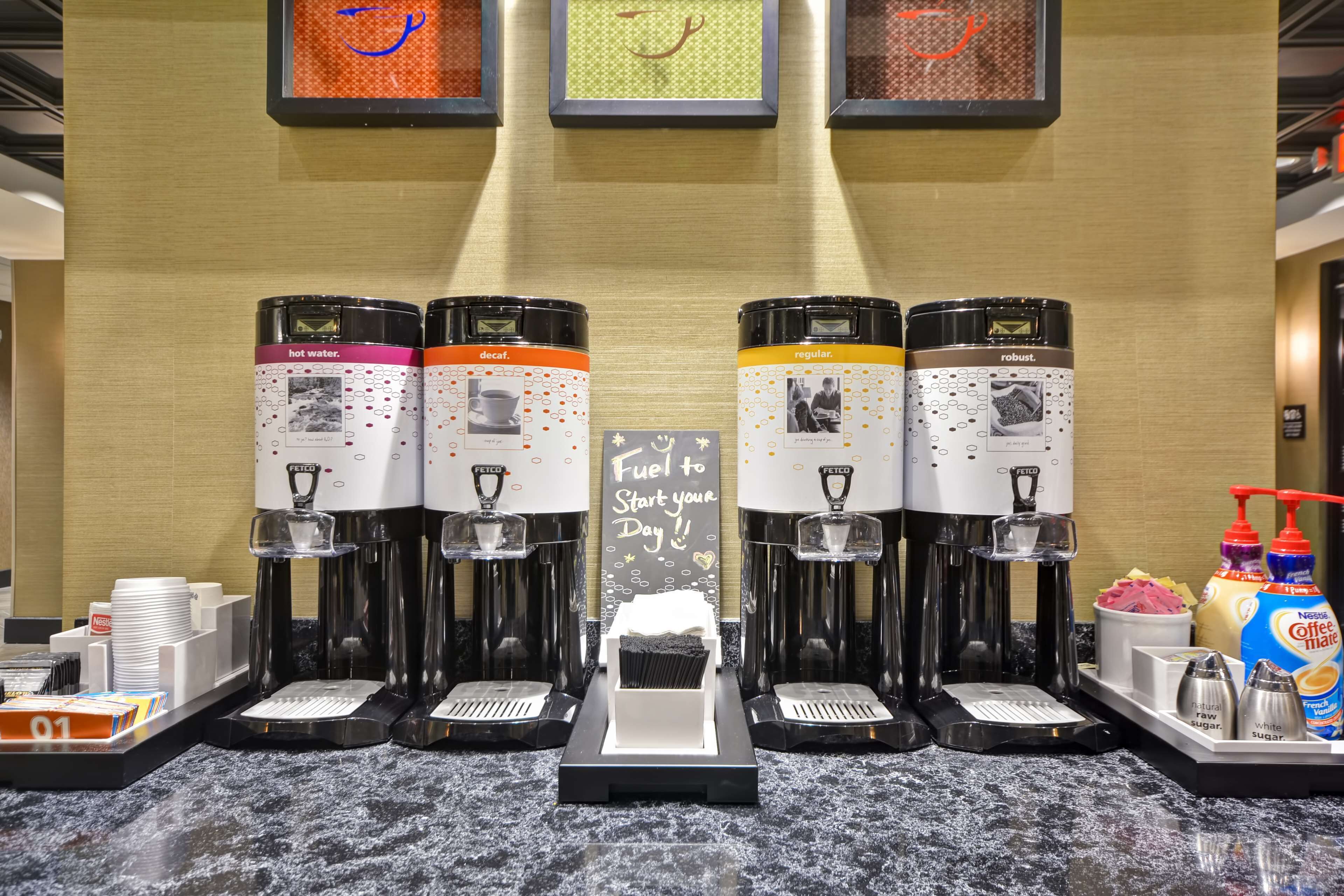 Vier koffieautomaatjes met verschillende labels voor heet water, gewone en sterke koffie, en een bord met de tekst 'Brandstof voor een Goede Start!'