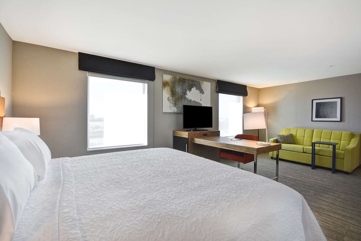 A hotel room with a large bed, a desk with a flat-screen TV, a green sofa, and a window providing a view of the city.