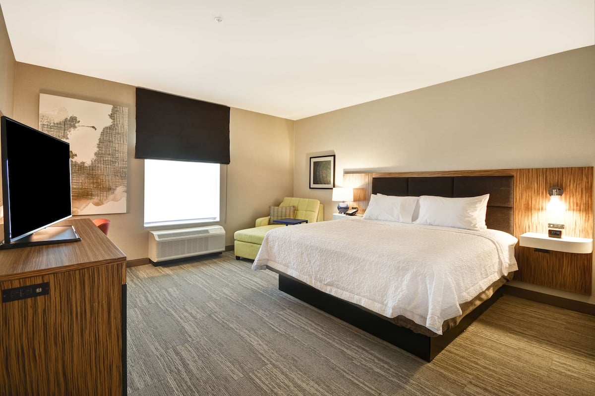 A hotel room with a large bed, a flat-screen TV, a desk, a chair, a window with blinds, and a wall-mounted lamp.